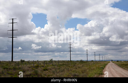 A long row of electricity transmission towers and high voltage power lines passing into the distant horizon, blue sky and clouds in a rural landscape Stock Photo