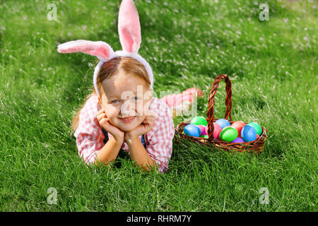 A cheerful little girl preschooler dressed in bunny ears is lying on the lawn with a basket of painted Easter eggs. Happy Easter Stock Photo