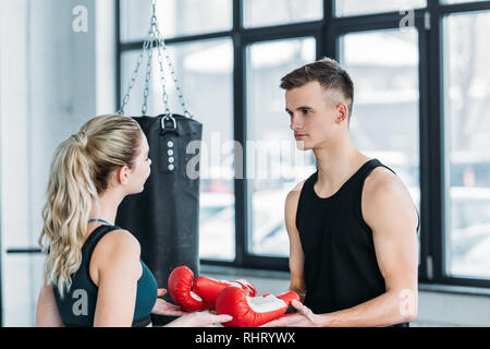 young male trainer giving boxing gloves to sporty young woman in gym Stock Photo