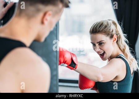 selective focus of male trainer looking at aggressive sportswoman boxing with punching bag in gym Stock Photo