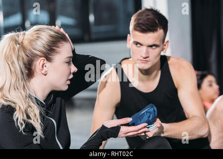 young sportsman giving ice pack to upset sporty girl suffering from head injury in gym Stock Photo