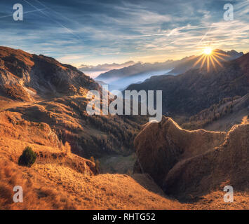 Mountains in fog at beautiful sunset in autumn. Dolomites, Italy. Landscape with alpine mountain valley, orange grass, low clouds, trees on hills, blu Stock Photo