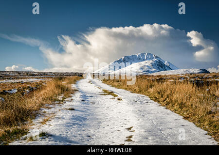 This is an remote dirt road covered in snow that disappears over the horizon.  In the distance a snow capped Errigal mountain can be seen surrounded b Stock Photo