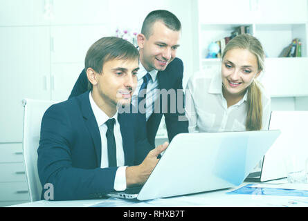 three friendly business male and female colleagues in formalwear working using laptops and talking in firm office Stock Photo