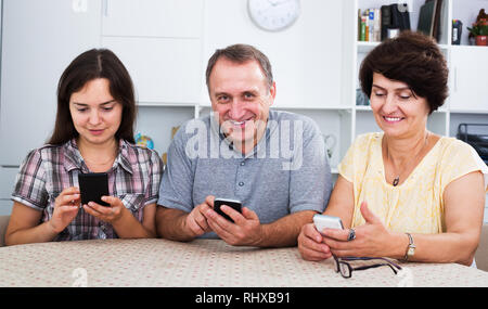 Positive senior couple and young woman using mobile phones while sitting together at home Stock Photo