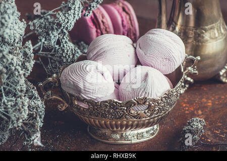 Freshly baked marshmallows and macaroons on metal plate with small white flowers and tea appliances composition on wooden background. Selective focus Stock Photo