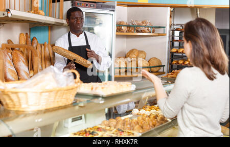 Polite male baker working behind counter in store of his bakery, serving female customer Stock Photo