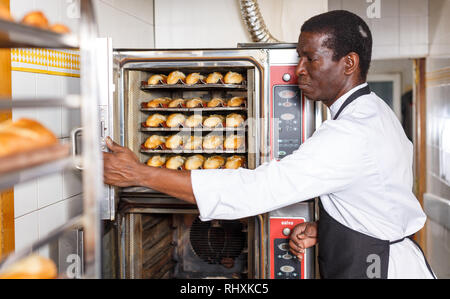 Portrait of African American baker controlling process of baking bread in professional oven at bakery Stock Photo