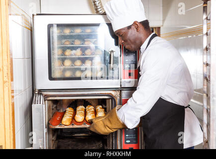 Experienced baker working in small bakery, taking out bread from industrial oven Stock Photo