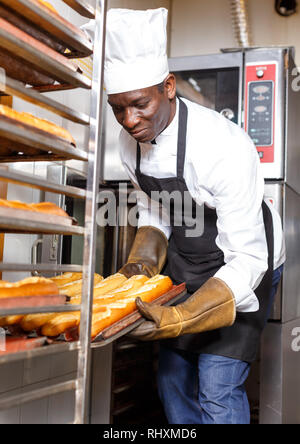 Experienced baker working in small bakery, taking out bread from industrial oven and putting on rack Stock Photo