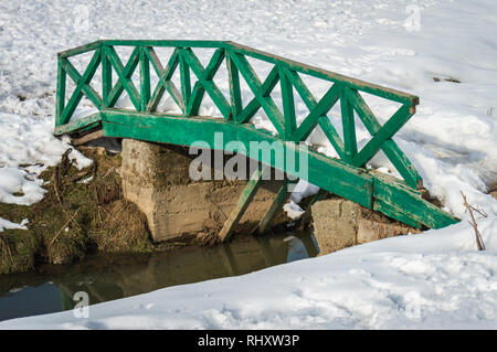 A green colour small wooden bridge over an small stream supported by concrete piers river in a snow landscape Stock Photo