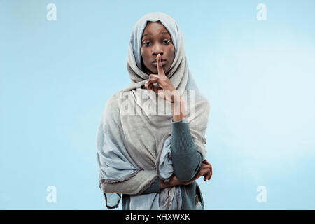 Secret, gossip concept. Young african woman whispering a secret behind her hand. The woman isolated on trendy blue studio background. Young emotional woman. Human emotions, facial expression concept. Stock Photo