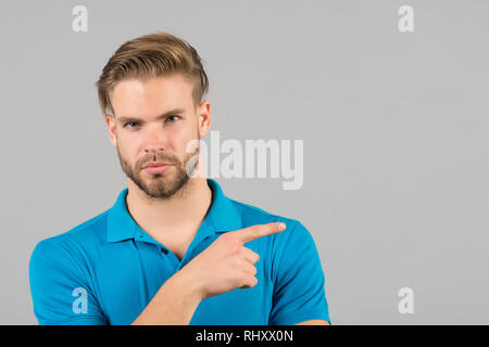 Advice advertisement concept. Guy bearded attractive pointing index finger to side. Man strict face looks confidently, grey background. Man with beard unshaven guy looks handsome well groomed. Stock Photo