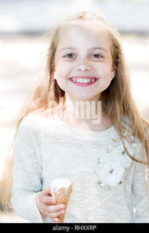 Girl sweet tooth on happy face eats ice cream, light background. Kid girl with ice cream cone in hand. Summer treats concept. Sweet tooth girl child with white ice cream in waffle cone. Stock Photo