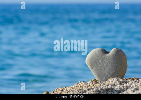 Big, grey heart shapped stone in a pebble on a beach against blurred sea and sky background, good as copy space Stock Photo