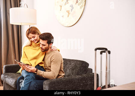 Hollidays planning. Young woman sitting at her boyfriends knees and dreaming. Happy mature couple with suitcases searching in tablet in hotel room Stock Photo