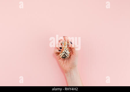 Creative top view flat lay of woman hand with black manicure holding small golden pineapple with copy space on pink paper background in minimalism sty Stock Photo