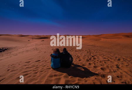 Night landscape with loving couple, boy and girl sitting on the sand dunes looking the horizon . Night sky with stars in the desert Sahara, Morocco Stock Photo