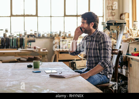 Woodworker sitting at a workbench talking on his cellphone Stock Photo