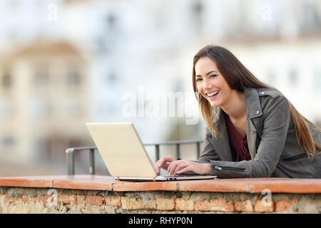 Happy woman using a laptop and looking at you in a balcony in a town outskirts Stock Photo