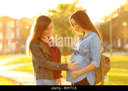 Happy pregnant woman showing belly to a friend in a park at sunset Stock Photo