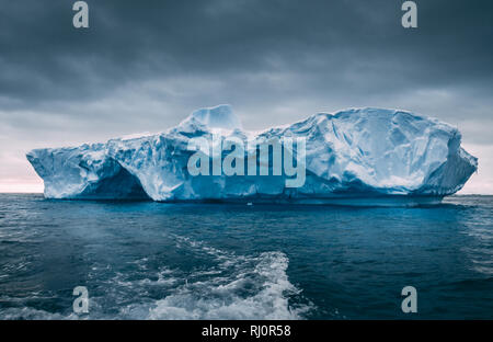 Big massive iceberg floating among the frozen ocean. Antarctica epic scenery in blue and grey tints. The dark cloudy sky over the South pole. The wild harsh environment. Mysterious winter landscape. Stock Photo