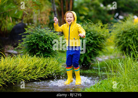 Kid playing in the rain in autumn park. Child jumping in muddy puddle on rainy fall day. Little boy in rain boots and yellow jacket outdoors in heavy  Stock Photo