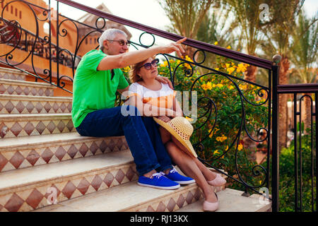 Senior couple sitting on stairs on hotel territory and pointing at view. People enjoying vacation. Traveling concept Stock Photo