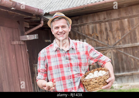 Smiling mature farmer holding fresh eggs in wicker basket at farm Stock Photo