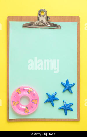 Flat lay of blank paper clipboard with sea stars and inflatable pool float on yellow background minimal summer vacation creative concept. Stock Photo