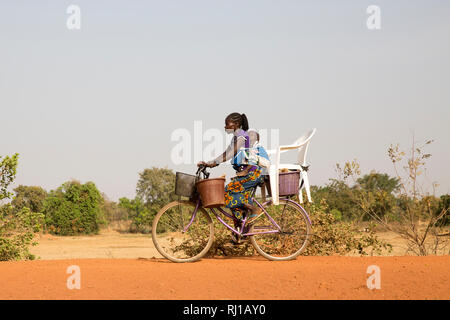 Samba village, Yako Province, Burkina Faso: A young woman carries her baby on her back and buckets and a chair on her bike. Stock Photo