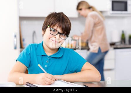 Positive schoolboy doing homework using laptop at kitchen interior, woman cooking at background Stock Photo
