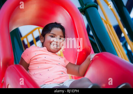 A happy and energetic girl playing on a red playground slide, about to slide down. Stock Photo