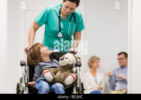 Smiling nurse pushing boy with teddybear on wheelchair while patients in hospital corridor Stock Photo