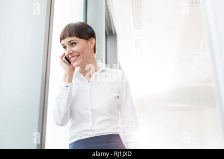 Smiling businesswoman talking on smartphone while standing by window at office Stock Photo