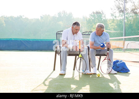 Full length of mature male friends talking while sitting with tennis rackets and balls on chair at court Stock Photo