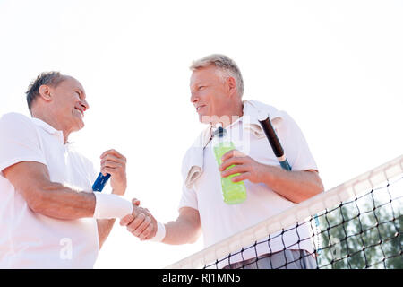 Low angle view of smiling men shaking hands while standing at tennis court against clear sky on sunny day Stock Photo