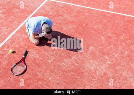 High angle view of disappointed mature man with head in hands while kneeling by tennis racket on red court during summer Stock Photo