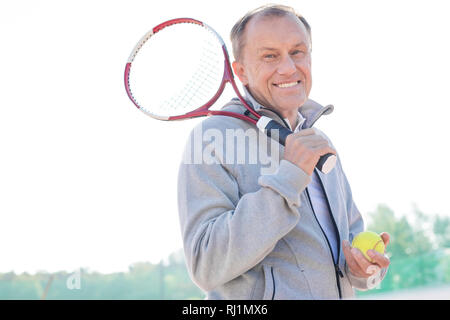 Portrait of smiling retired senior man standing with tennis racket and ball against clear sky on sunny day Stock Photo