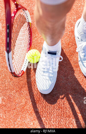 Low section of senior man standing with tennis racket and ball on red court Stock Photo