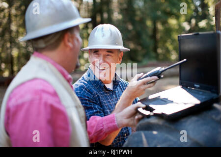 Two lumberjacks in the forest, one on his walkie talkie, the other using his laptop. Stock Photo