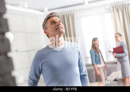 Mature man looking up while standing against saleswoman talking with woman in apartment Stock Photo