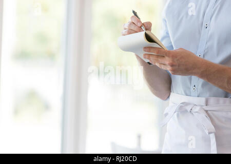 Midsection of young waiter writing on notepad while standing at restaurant Stock Photo