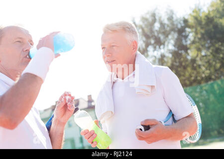 Mature man looking senior friend drinking from bottle while standing on tennis court during summer Stock Photo