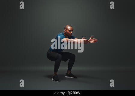 Full length of strong young muscular man in comfortable sportwear stretching his body. Fitness african male model standing over dark background Stock Photo