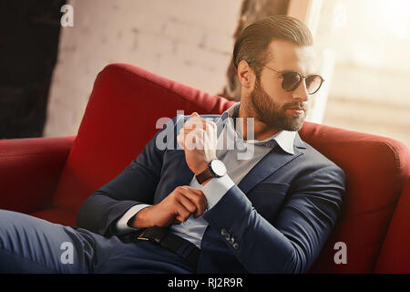 Young and successful...Businessman is sitting on sofa in modern office and looking away through sunglasses. Fashion look. Business style. Stock Photo