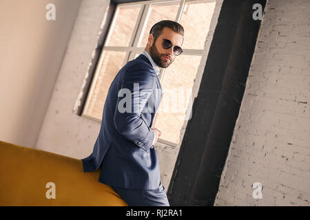 Being successful. Portrait of pleasant and young businessman in sunglasses and stylish suit who is standing at office. Business look. Close-up image. Stock Photo