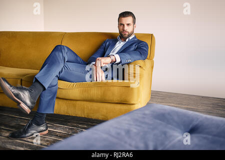 Good looking young businessman in a stylish suit is relaxing on sofa and looking at camera. Business look. Stock Photo