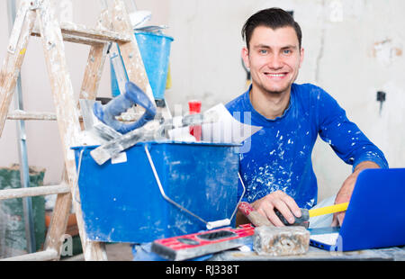 Smiling happy male contractor working with papers and laptop in repairable apartment Stock Photo