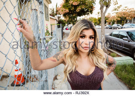 Portrait confident, tough Latinx young woman with tattoos leaning against neighborhood fence
